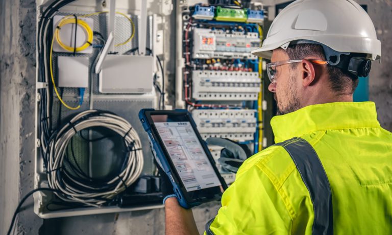 Man, an electrical technician working in a switchboard with fuses, uses a tablet.